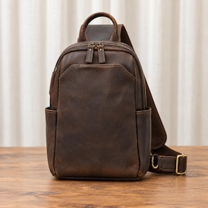 Könnte beinhalten: Ein dunkelbrauner Leder-Sling-Rucksack mit einem einzelnen Schulterriemen, einem Tragegriff und mehreren Reißverschlussfächern. Die Tasche hat eine glatte, matte Oberfläche mit sichtbaren Nähten, Seitentaschen und Beschlägen aus antikem Messing.