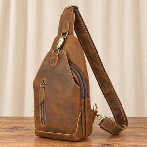 Pode incluir: Uma bolsa tiracolo de couro castanho com um bolso frontal vertical com fecho, um compartimento principal com fecho duplo e uma alça ajustável fixada com um clipe giratório de metal e detalhes em latão.