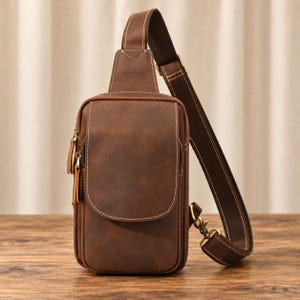 Pode incluir: Uma bolsa tiracolo de couro castanho com formato retangular vertical, apresentando um bolso frontal com aba e costuras visíveis, além de uma alça ajustável com ferragens metálicas. A bolsa possui um acabamento liso e fosco.