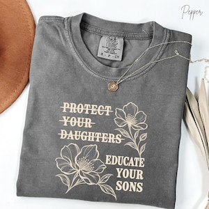 May include: A pepper-colored short-sleeve t-shirt featuring the text "PROTECT YOUR DAUGHTERS" with lines through the words and "EDUCATE YOUR SONS" in a light cream color. The shirt includes two line-art floral illustrations and a neck label.