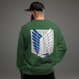 Könnte beinhalten: Ein waldgrünes Langarm-Sweatshirt mit einer großen Grafik auf der Rückseite. Das Design zeigt das Emblem der Flügel der Freiheit, bestehend aus zwei stilisierten Flügeln, einem blauen und einem weißen, vor einem hellgrauen Schildhintergrund.