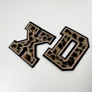 K&ouml;nnte beinhalten: Zwei Buchstaben-Aufn&auml;her im College-Stil, die Buchstaben X und D, mit einem braun-schwarzen Leopardenmuster. Jeder Buchstabe ist mit einem d&uuml;nnen metallisch goldenen Rand und einer dicken schwarzen Au&szlig;enkante versehen, was f&uuml;r einen markanten Look sorgt.