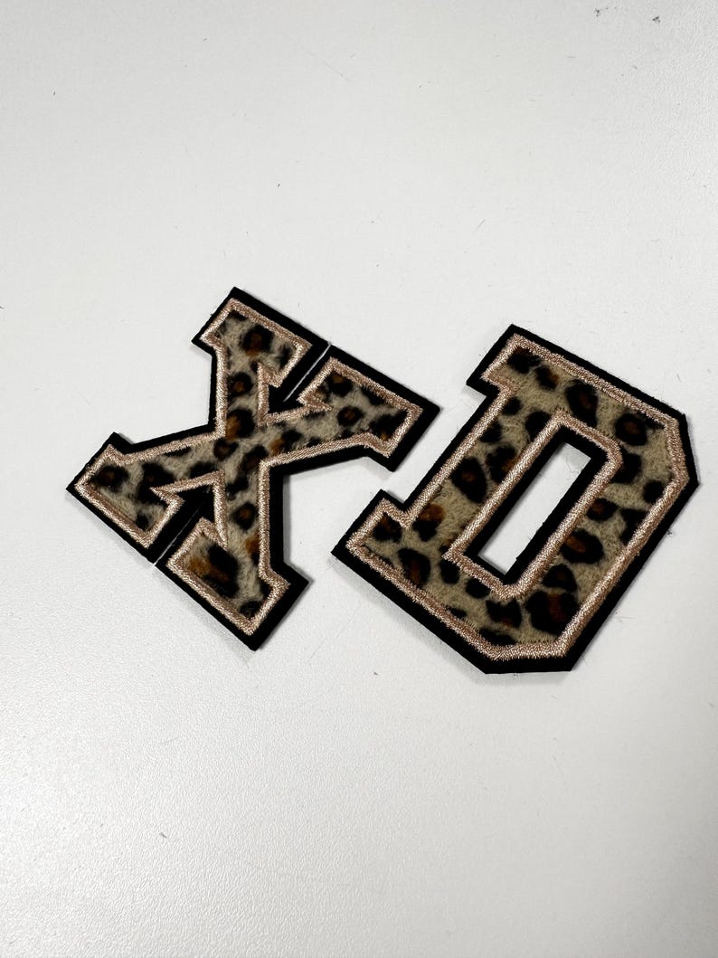 K&ouml;nnte beinhalten: Zwei Buchstaben-Aufn&auml;her im College-Stil, die Buchstaben X und D, mit einem braun-schwarzen Leopardenmuster. Jeder Buchstabe ist mit einem d&uuml;nnen metallisch goldenen Rand und einer dicken schwarzen Au&szlig;enkante versehen, was f&uuml;r einen markanten Look sorgt.