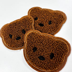 Könnte beinhalten: Drei braune, bärenförmige Aufnäher mit einer strukturierten, flauschigen Oberfläche. Jeder Aufnäher weist gestickte schwarze Faden-Details auf, die zwei runde Augen und eine kleine ovale Nase bilden, mit einem gestickten Rand.