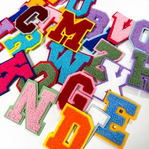 Puede incluir: Una colección de parches de letras estilo universitario con una superficie de felpilla texturizada y difusa. Cada letra está delineada con un borde de fieltro en contraste en varios colores vibrantes, incluyendo rojo, azul, verde, rosa, naranja, amarillo y morado.