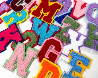 Könnte beinhalten: Eine Sammlung verschiedener Buchstaben-Aufnäher im College-Stil mit einer strukturierten, flauschigen Chenille-Oberfläche. Jeder Buchstabe ist mit einem kontrastierenden Filzrand in verschiedenen leuchtenden Farben wie Rot, Blau, Grün, Pink, Orange, Gelb und Lila versehen.