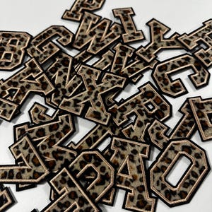 Könnte beinhalten: Eine Ansammlung verstreuter gestickter Buchstabenaufnäher mit braun-schwarzem Leopardenmuster. Jeder Buchstabe hat einen dicken schwarzen Rand und eine dünne metallisch goldene Umrandung im klassischen Varsity-Blockschriftstil.