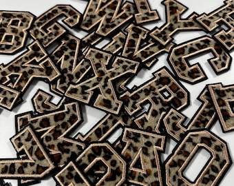 Könnte beinhalten: Eine Ansammlung verstreuter gestickter Buchstabenaufnäher mit braun-schwarzem Leopardenmuster. Jeder Buchstabe hat einen dicken schwarzen Rand und eine dünne metallisch goldene Umrandung im klassischen Varsity-Blockschriftstil.