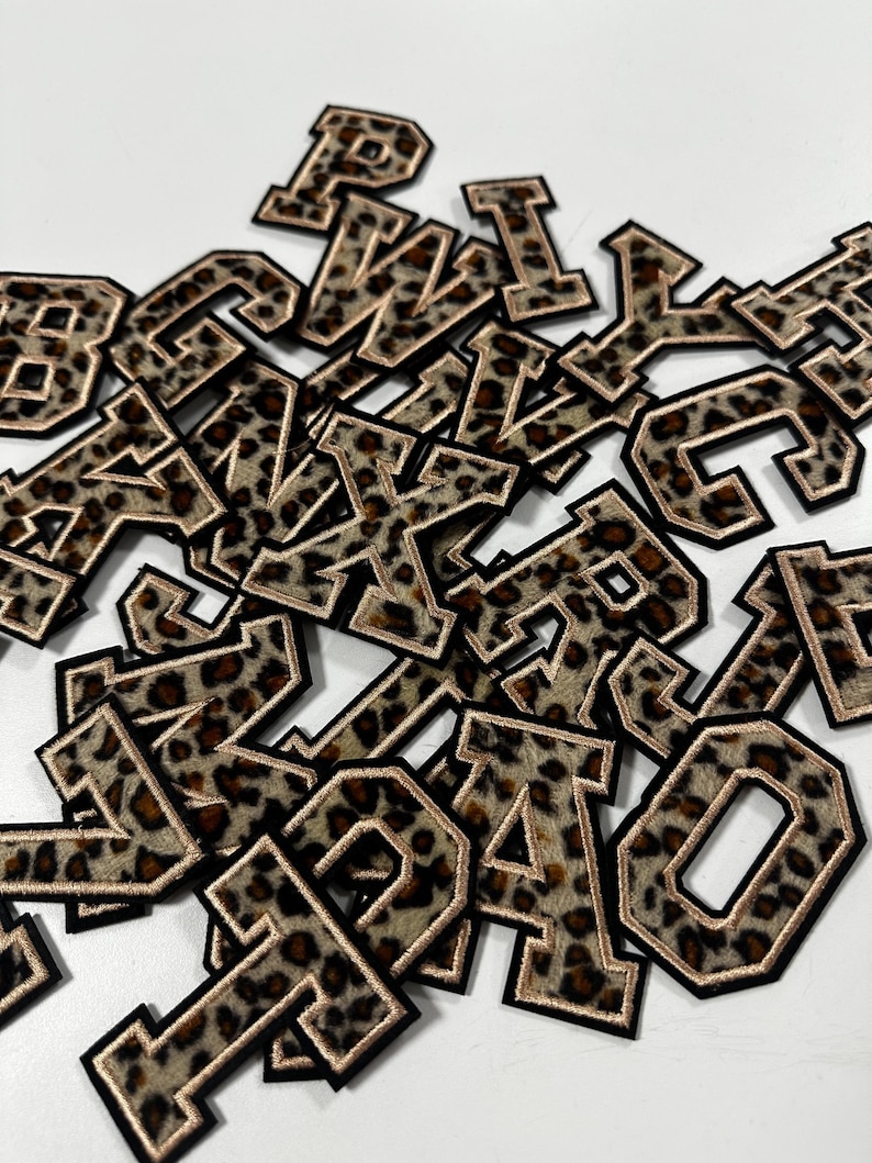 K&ouml;nnte beinhalten: Eine Ansammlung verstreuter gestickter Buchstabenaufn&auml;her mit braun-schwarzem Leopardenmuster. Jeder Buchstabe hat einen dicken schwarzen Rand und eine d&uuml;nne metallisch goldene Umrandung im klassischen Varsity-Blockschriftstil.