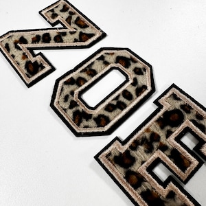 K&ouml;nnte beinhalten: Drei gestickte Buchstaben im College-Stil, die das Wort ZOE bilden, mit einem flauschigen Leopardenmuster in Braun-, Schwarz- und Cremet&ouml;nen, umrandet mit einem metallisch goldenen Faden und einer dicken schwarzen Filzkante f&uuml;r eine Textur.