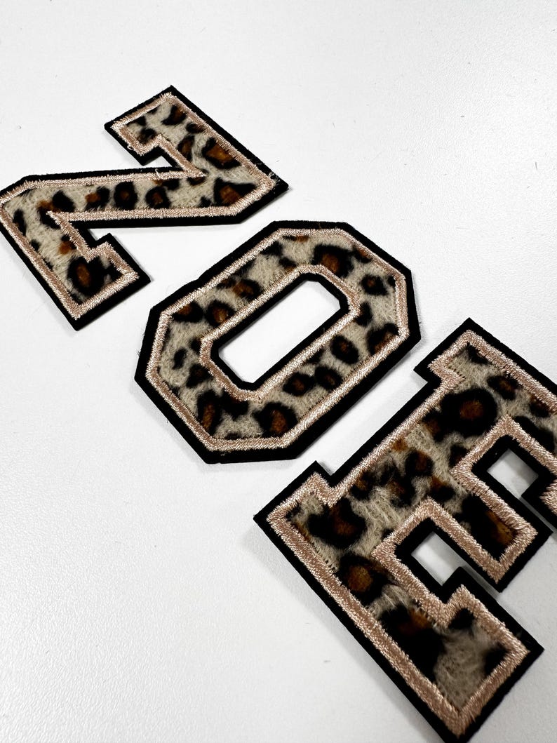 K&ouml;nnte beinhalten: Drei gestickte Buchstaben im College-Stil, die das Wort ZOE bilden, mit einem flauschigen Leopardenmuster in Braun-, Schwarz- und Cremet&ouml;nen, umrandet mit einem metallisch goldenen Faden und einer dicken schwarzen Filzkante f&uuml;r eine Textur.