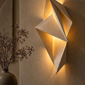 Op de afbeelding: Een geometrische wandlamp met een getextureerde gebroken witte afwerking, voorzien van scherpe hoekige vlakken die van binnenuit een warme, zachte gloed uitstralen. De lamp is op een vlak oppervlak gemonteerd en werpt licht naar boven en beneden.