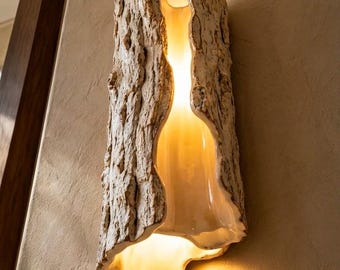 May include: A wall-mounted light fixture featuring a textured, bark-like exterior in shades of beige and cream. The vertical, cylindrical form is split down the center, revealing a smooth, glossy interior that emits a warm, soft, golden glow.