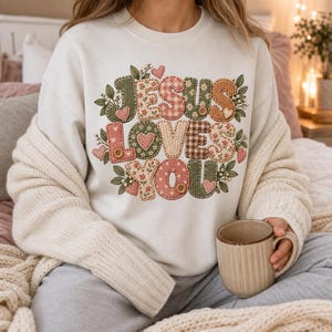 May include: A cream-colored crewneck sweatshirt featuring a graphic design with the text "JESUS LOVES YOU" in a patchwork style. The letters are filled with various patterns including floral, gingham, and polka dots in shades of green, pink, and brown.