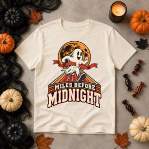 Puede incluir: Una camiseta de color crema con un gráfico de un fantasma de dibujos animados corriendo a través de una cinta de meta roja frente a una gran luna naranja. Debajo del fantasma, el texto "MILES BEFORE MIDNIGHT" aparece en letras oscuras.