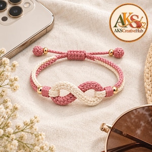 May include: A woven cord bracelet featuring an infinity symbol design in alternating dusty rose and cream colors. The bracelet includes small gold-toned metallic beads and an adjustable sliding knot closure. The logo AKS CreativeHub is visible.