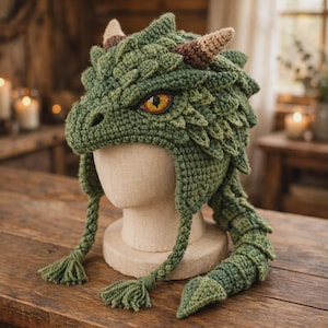 May include: A green crocheted dragon hat featuring textured scales, two small brown horns, and a single yellow eye with a black pupil. The hat includes two braided earflaps with tassels and a long, scaled tail extension hanging from the side.
