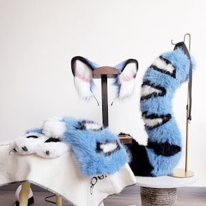 Op de afbeelding: Een set van imitatiebont accessoires bestaande uit een paar dierenoren op een haarband, een paar pootvormige handschoenen en een lange, gebogen staart. De items zijn lichtblauw, wit en zwart met een gestreept patroon op de staart en oren.