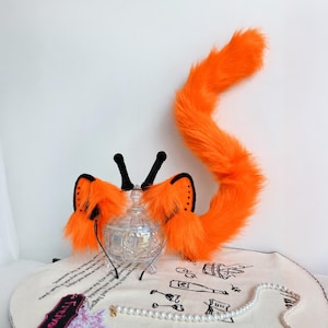 May include: A set of bright orange faux fur animal ears attached to a thin black headband, accompanied by a long, matching orange faux fur tail. The ears feature black inner detailing and black upright tufts at the top of each ear.