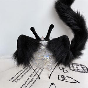 Puede incluir: Una diadema de piel sintética negra con dos orejas de animal erguidas y dos tallos tipo antena, acompañada de una larga cola de piel sintética negra a juego. El conjunto se exhibe sobre un recipiente de vidrio transparente y facetado.
