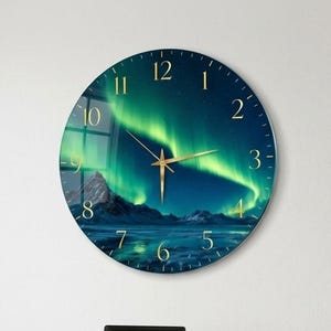 Pode incluir: Um relógio de parede circular com uma paisagem de aurora boreal em tons de verde e azul sobre uma cordilheira nevada. O mostrador apresenta numerais dourados e ponteiros finos de cor dourada que contrastam com o céu noturno escuro.