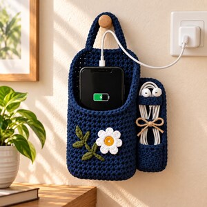 May include: A dark blue crocheted wall hanging organiser featuring a daisy applique with green leaves. The main pocket holds a black smartphone charging with a white cable, while a smaller side pocket stores white wired earbuds with a tan cord tie.
