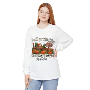May include: A white long-sleeve shirt featuring a graphic of a farm scene with pumpkins, a house, a windmill, and a tree. The text reads I will praise Him in every season Psalm 34:1 in black script and block lettering across the chest area.