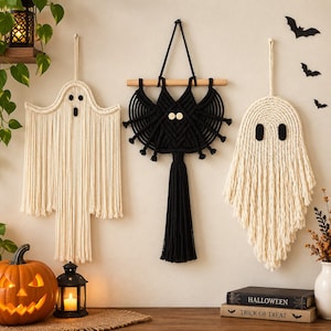 May include: Three macrame wall hangings featuring two off-white ghosts with black felt eyes and one black bat with two light-colored circular buttons for eyes. The bat is suspended from a wooden dowel, while the ghosts hang from loops.