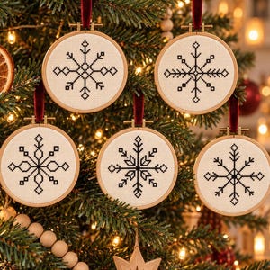 May include: Five circular wooden embroidery hoops containing white fabric with black cross-stitched snowflake patterns. Each ornament hangs from a dark red velvet ribbon, displaying a unique geometric snowflake design centered on the fabric.