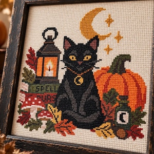 Könnte beinhalten: Ein gerahmtes Kreuzstich-Stickbild mit einer schwarzen Katze mit gelben Augen und Mondanhänger, einer leuchtenden Laterne, einem großen orangefarbenen Kürbis, roten Pilzen, Herbstblättern sowie einem gelben Halbmond mit Sternen auf beigem Stoff.