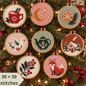 May include: Eight circular wooden embroidery hoops featuring cross-stitch designs of a red mug, a crescent moon with stars, an orange slice, a white bird, a dark green teapot, a house in the snow, holly berries, and a fox, with 39 x 39 stitches.