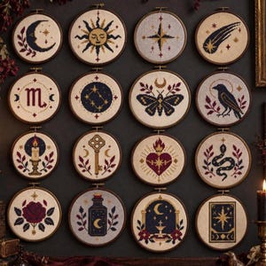 Puede incluir: Una colección de dieciséis bastidores de bordado circulares con diseños de punto de cruz que incluyen una luna creciente, sol, estrella, cometa, símbolo de escorpión, constelación, polilla, cuervo, vela, llave, corazón sagrado, serpiente, rosa, frasco, arco y carta del tarot.