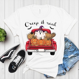 May include: A white t-shirt featuring a graphic of a red pickup truck carrying three jack-o'-lanterns, hay bales, and a ghost wearing a purple witch hat. The text "Creep it real" is printed above the truck, accompanied by two black bats.
