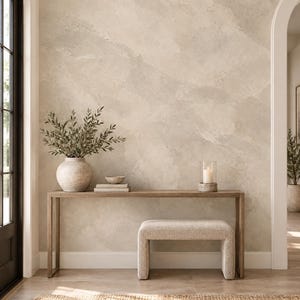 May include: A rectangular wooden console table with thin legs, featuring a textured beige vase with green branches, a small bowl, stacked books, and a glass candle holder. A beige upholstered bench sits underneath the table.