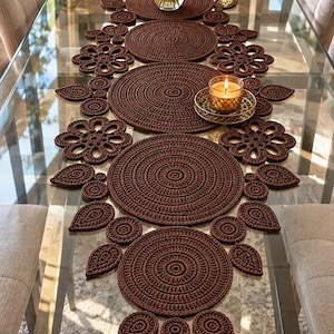 May include: A dark brown crochet table runner featuring a series of large circular mats connected by smaller circular and leaf-shaped motifs. The runner is arranged down the center of a glass table, accented by a glass vase and a candle.