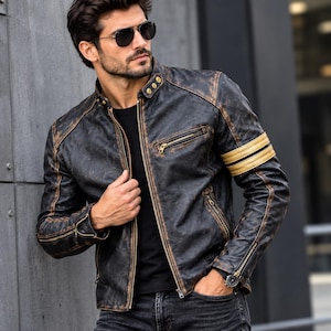 Può includere: Una giacca in pelle nera dall'aspetto vissuto con colletto alla coreana, cerniere in metallo color oro e due strisce orizzontali beige sulla manica sinistra. La giacca presenta una chiusura frontale con zip, bottoni a pressione e polsini con zip.