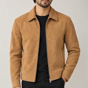 May include: A tan suede jacket featuring a classic collar, a full-length silver-toned metal zipper, long sleeves with buttoned cuffs, and two horizontal seam details across the chest, worn over a plain black crew neck t-shirt.