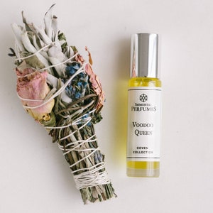 May include: A bundle of dried herbs and flowers tied with twine, next to a perfume bottle. The bottle has a silver cap and a label that reads "Voodoo Queen" from the "Coven Collection" by Immortal Perfumes.