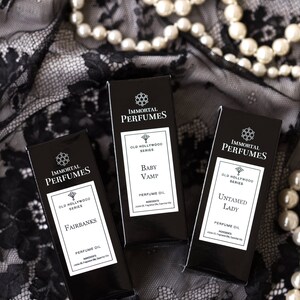 May include: Three black perfume bottles with gold lettering. The bottles are labeled "Immortal Perfumes" and feature the names "Fairbanks", "Baby Vamp", and "Untamed Lady". Each bottle is labeled "Old Hollywood Series" and "Perfume Oil".
