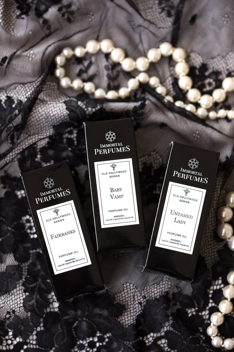 May include: Three black perfume bottles with gold lettering. The bottles are labeled "Immortal Perfumes" and feature the names "Fairbanks", "Baby Vamp", and "Untamed Lady". Each bottle is labeled "Old Hollywood Series" and "Perfume Oil".