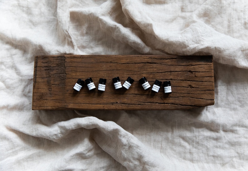 May include: Ten small black bottles with white labels on a rustic wooden board. The labels read "Amber & Lavender Essential Perfume".