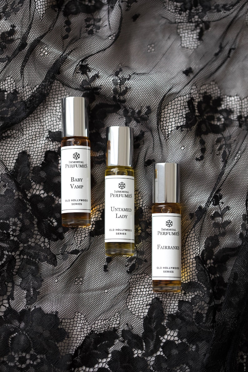 May include: Three small glass bottles of perfume with silver caps and labels. The labels are white with black text and feature the brand name "Immortal Perfumes" and the names of the scents: "Baby Vamp", "Untamed Lady", and "Fairbanks". All three bottles are part of the "Old Hollywood" series.