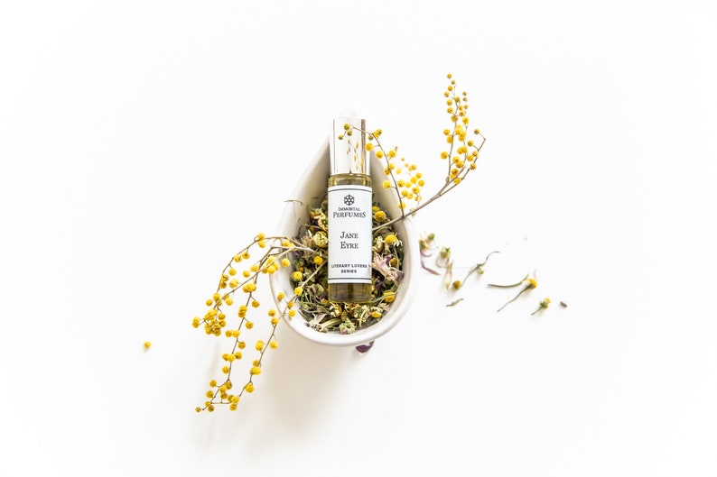 May include: A small glass bottle of perfume with a white label that reads "Jane Eyre" and "Library Lotion Series". The bottle is sitting in a white ceramic bowl filled with dried flowers and yellow flower sprigs.