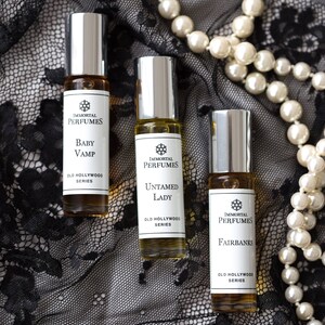 May include: Three small glass bottles of perfume with silver caps and labels that say "Immortal Perfumes" and "Old Hollywood Series". The bottles are on a black lace fabric background with a pearl necklace.