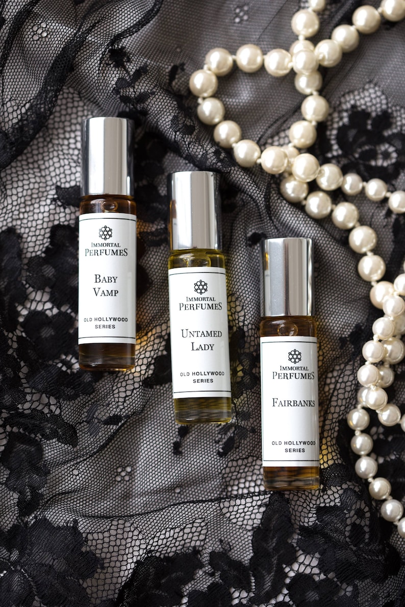 May include: Three small glass bottles of perfume with silver caps and labels that say "Immortal Perfumes" and "Old Hollywood Series". The bottles are on a black lace fabric background with a pearl necklace.