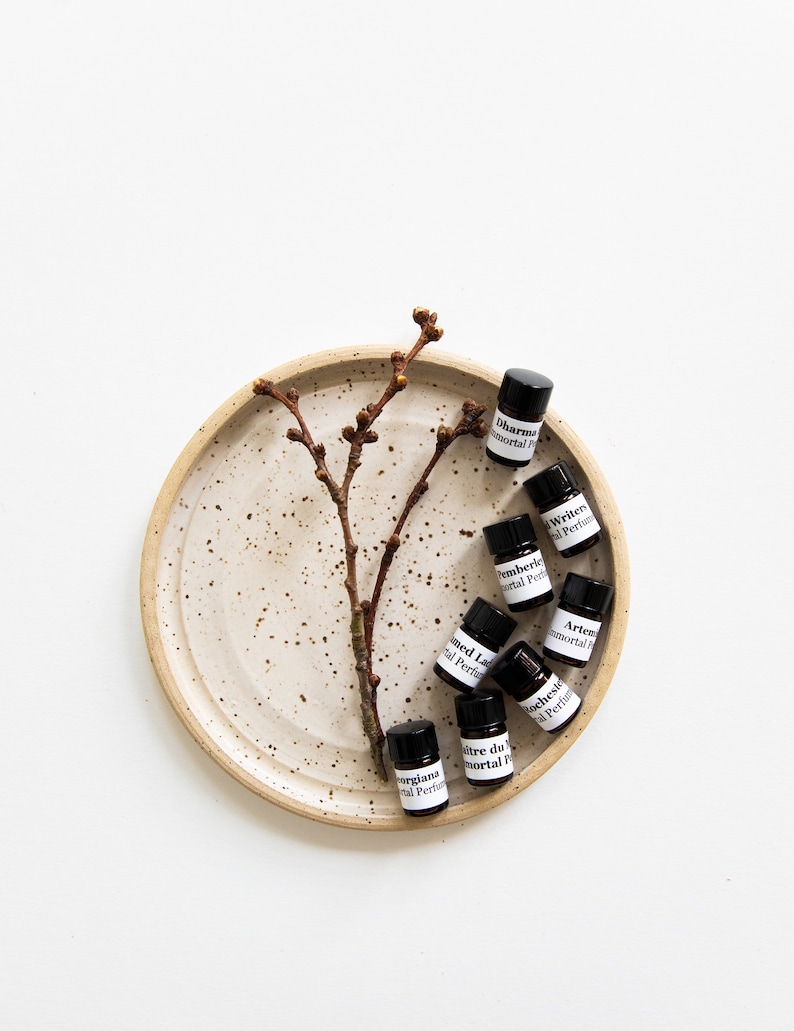 May include: A collection of small, dark glass bottles with white labels containing essential oils. The bottles are arranged on a speckled, beige ceramic plate with a small, brown twig branch.