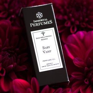 May include: A black bottle of perfume oil labeled "Immortal Perfumes Old Hollywood Series Baby Vamp Perfume Oil". The bottle is surrounded by dark red flowers.