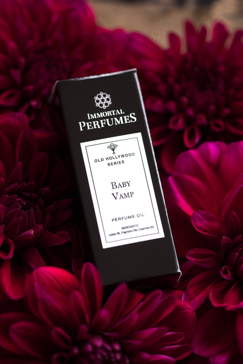 May include: A black bottle of perfume oil labeled "Immortal Perfumes Old Hollywood Series Baby Vamp Perfume Oil". The bottle is surrounded by dark red flowers.