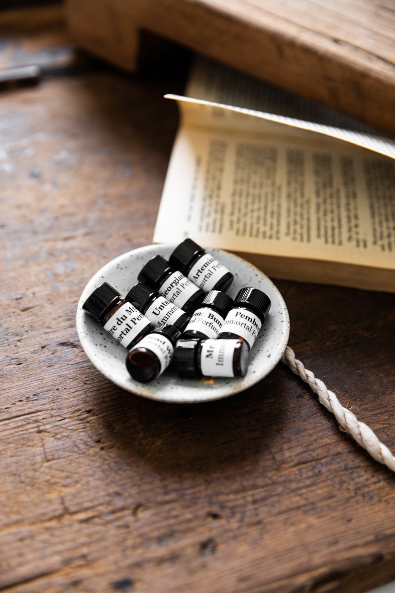 May include: A small white ceramic dish with eight small glass bottles containing essential oils. The bottles are labeled with the names of the scents, including "Tre du My", "Georgian", "Untaortal", "Artemis", "Pember", "Mr", "Imm", and "Bun".