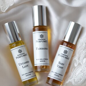 May include: Three small glass bottles of perfume with silver caps. The bottles are labeled with the text "Immortal Perfumes" and the names of the scents: "Fairbanks", "Untamed Lady", and "Baby Vamp". Each bottle is labeled with "Old Hollywood Series".